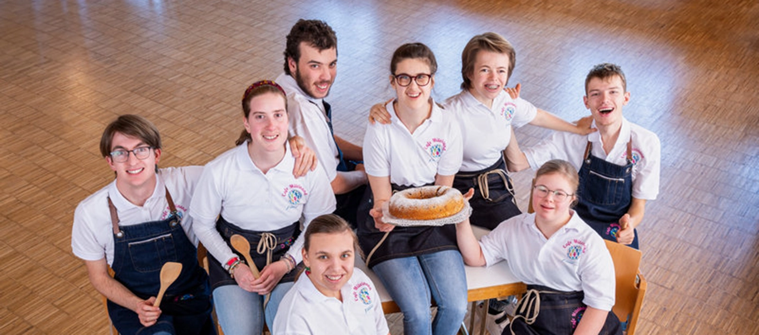 Teamfoto Cafe Miteinand ausgestattet in Firmenkleidung und mit Kochlöffeln und einem Kuchen